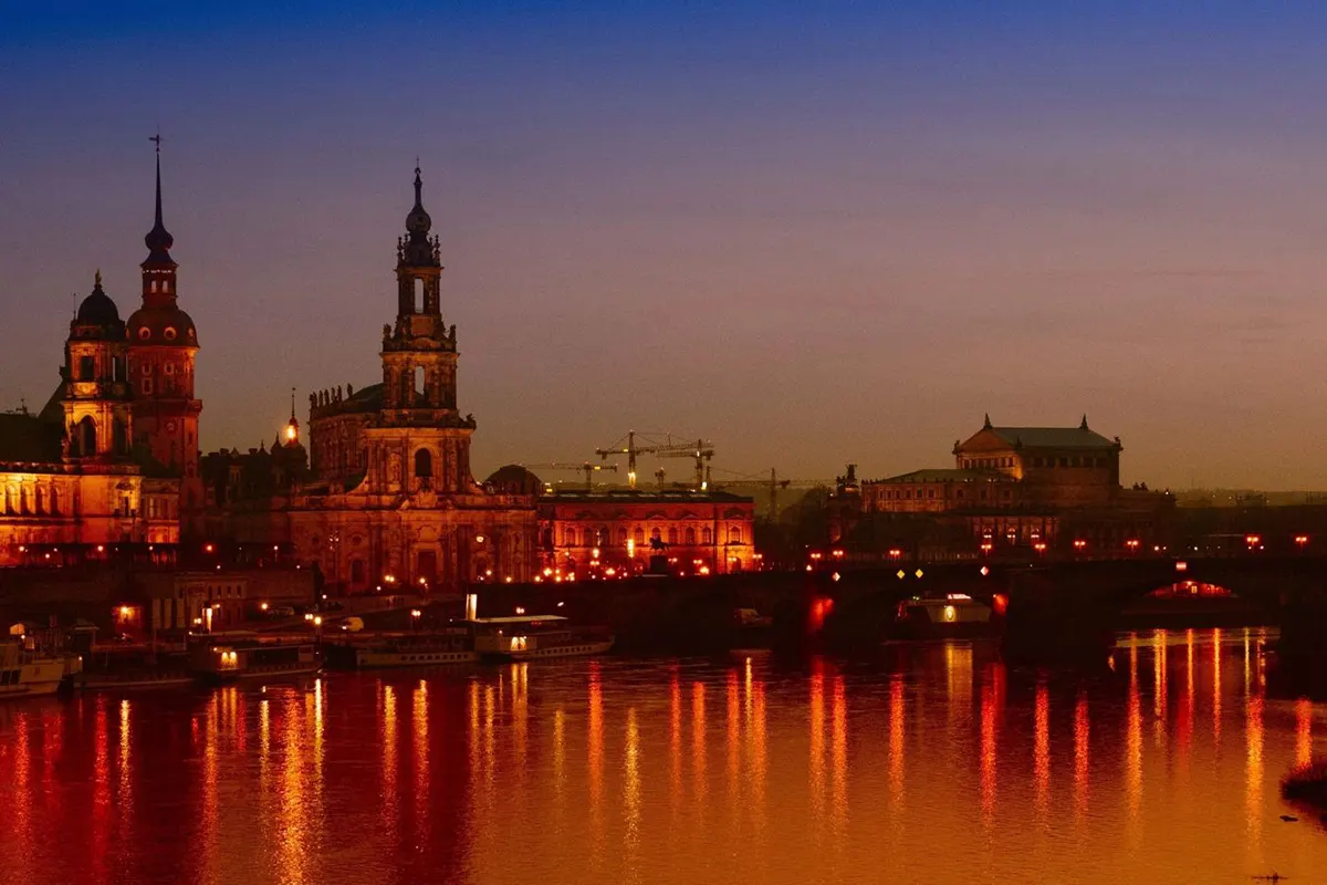 Die Altstadt von Dresden bei Dämmerung: Historische Gebäude wie Frauenkirche und Semperoper leuchten warm, ihre Lichter spiegeln sich in der Elbe.