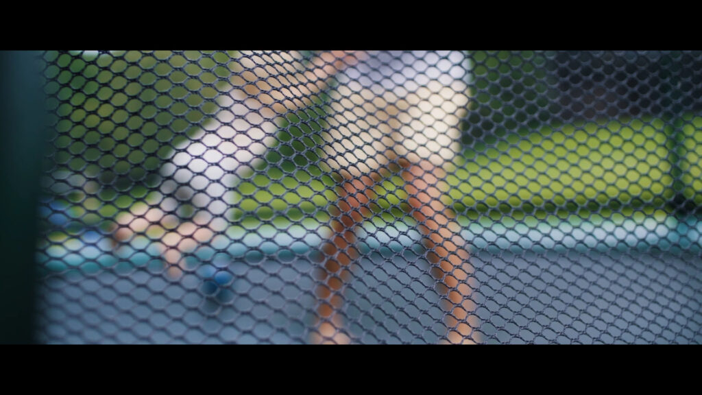 Eine erwachsene Person spielt mit einem Kind auf einem Trampolin. Gefilmt durch das Sicherheitsnetz des Trampolins. Die Personen sind unscharf. Keine Gesichter zu erkennen.