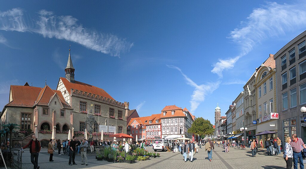 Blick auf den historischen Marktplatz von Göttingen mit dem markanten Alten Rathaus auf der linken Seite, das eine charakteristische Fassade mit rotem Ziegeldach und Turm aufweist. Die Fußgängerzone ist belebt mit Menschen, umgeben von farbenfrohen Fachwerk- und Bürgerhäusern. Im Hintergrund sind weitere historische Gebäude und Kirchtürme unter einem strahlend blauen Himmel mit zarten Schleierwolken zu sehen.