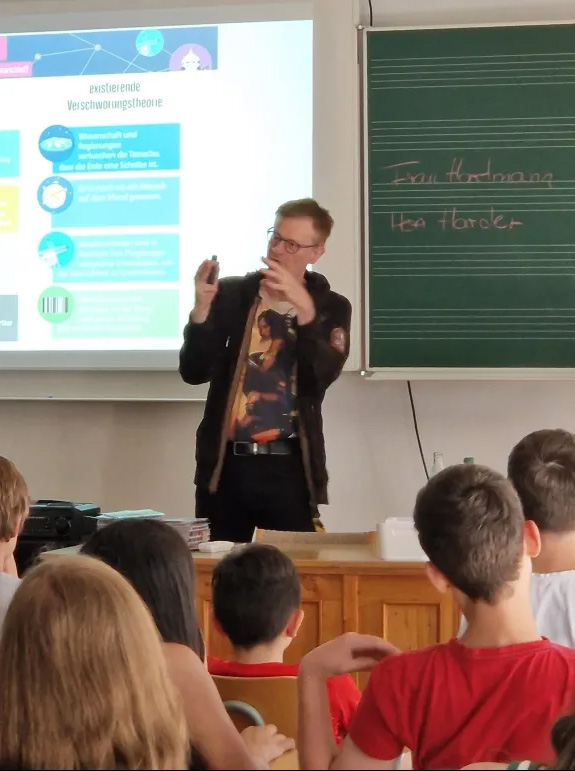 Bernd Harder bei Veranstaltungen mit Schulklassen, während er gerade etwas zum Thema Verschwörungstheorie erklärt. Auf der Leinwand im Hintergrund steht als Überschrift "existierende Verschwörungstheorie".