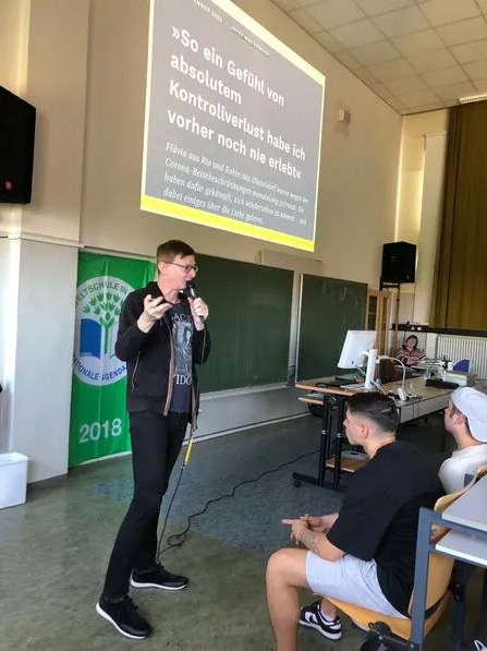 Bernd Harder bei Veranstaltungen mit Schulklassen, während er gerade etwas zum Thema Verschwörungstheorie erklärt. Auf der Leinwand ist ein Zitat zu lesen: "So ein Gefühl von absolutem Kontrollverlust habe ich vorher noch nie erlebt."