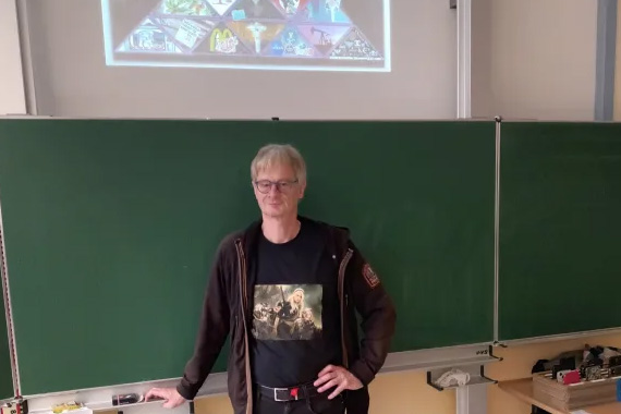 Bernd Harder steht im Klassenraum vor einer Tafel. Im Hintergrund sieht man auf einem Beamer Symbole zu Verschwörungstheorien in einer Pyramide