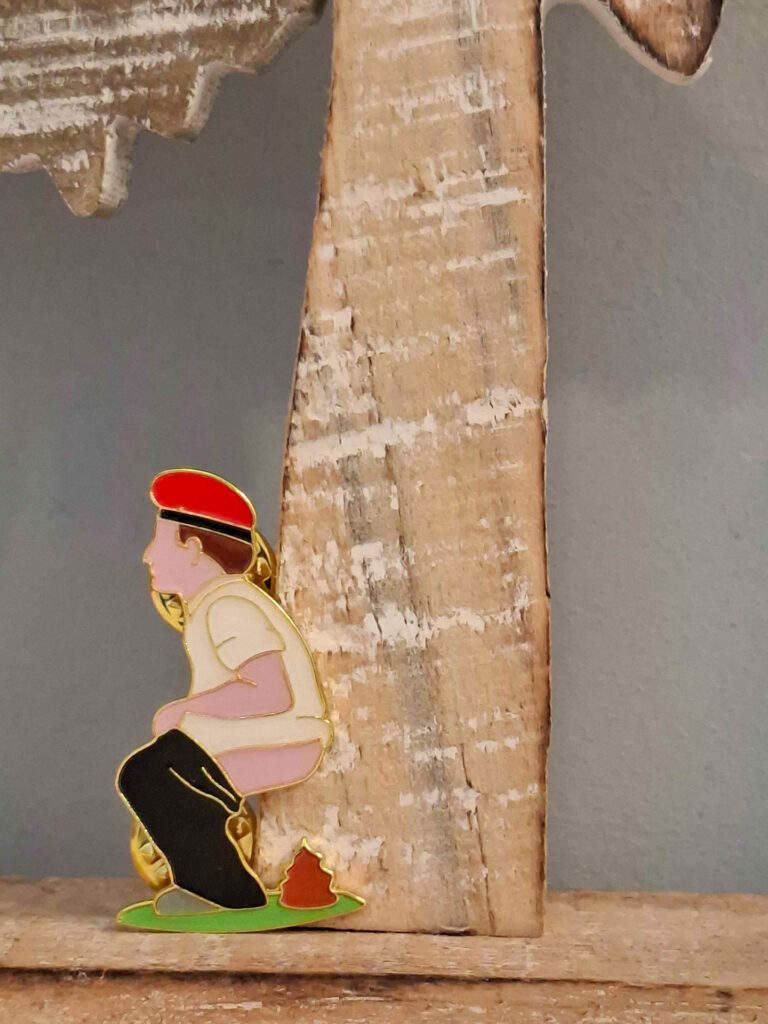 Vor einem geschnitzten Holzbaum hockt eine Figur. Ein Mann mit einer roten Mütze und einem weißen T-Shirt hockt, die schwarze Hose heruntergelassen am Stamm. Unter seinem Po liegt ein braunes Häufchen.