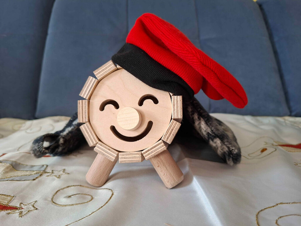 Auf einem blauen Sofa steht ein kurzer Baumstamm mit eingeschnitztem Gesicht. Er trägt eine rote Mütze und ist mit einer Flanell-Decke zugedeckt.