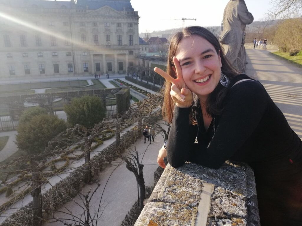 Diana Becht lächelt in die Kamera und zeigt ein Peace-Zeichen vor einer historischen Garten- und Schlossanlage im Sonnenlicht.