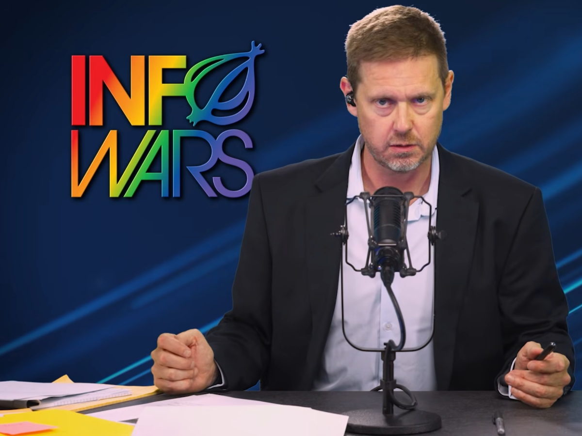 Tim Heidecker, ein mittelalter Mann im Anzug sitzt an einem Schreibtisch vor einem Mikrofon. Im Hintergrund ein regenbogenfarbenes "InfoWars" Logo mit einer stilisierten Zwiebel als O.